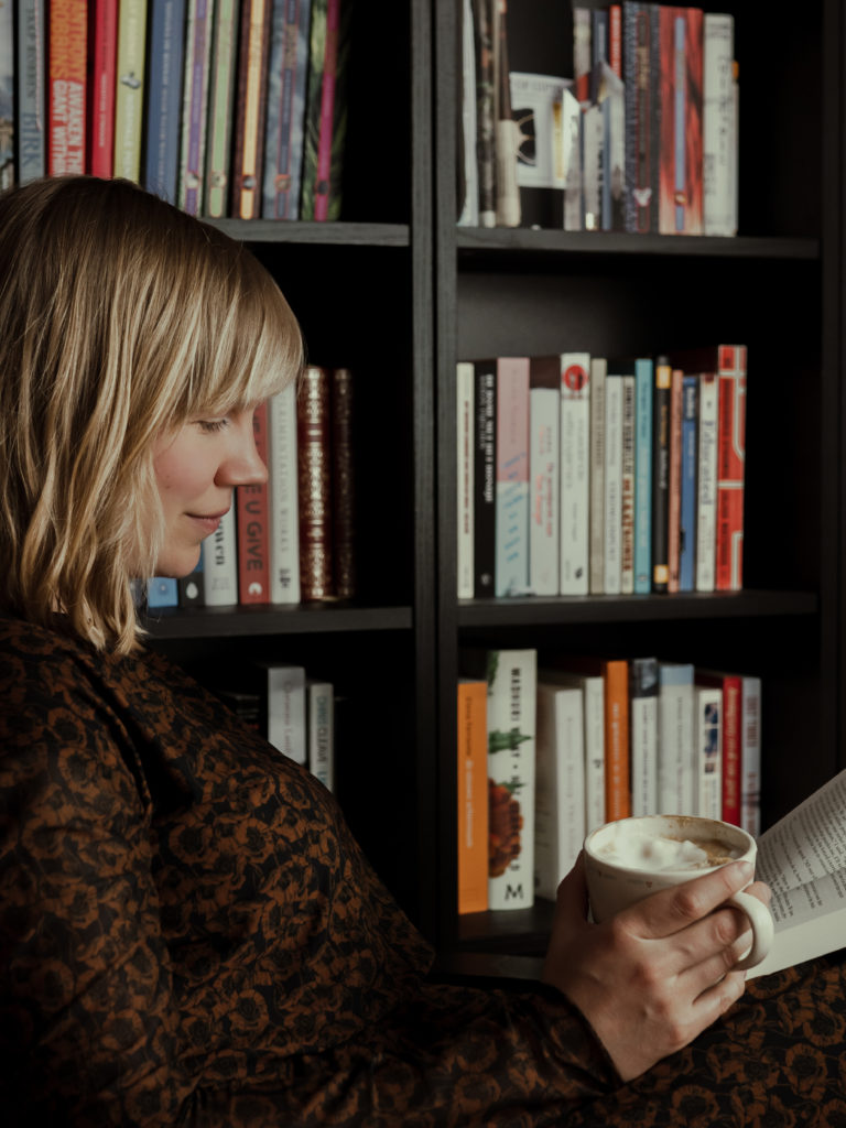Lucia van den Brink leest een boek en drinkt koffie naast een boekenkast. Ze is schrijver, literair talent, schrijft over trauma en jeugdtrauma en (emotionele) mishandeling en verwerking